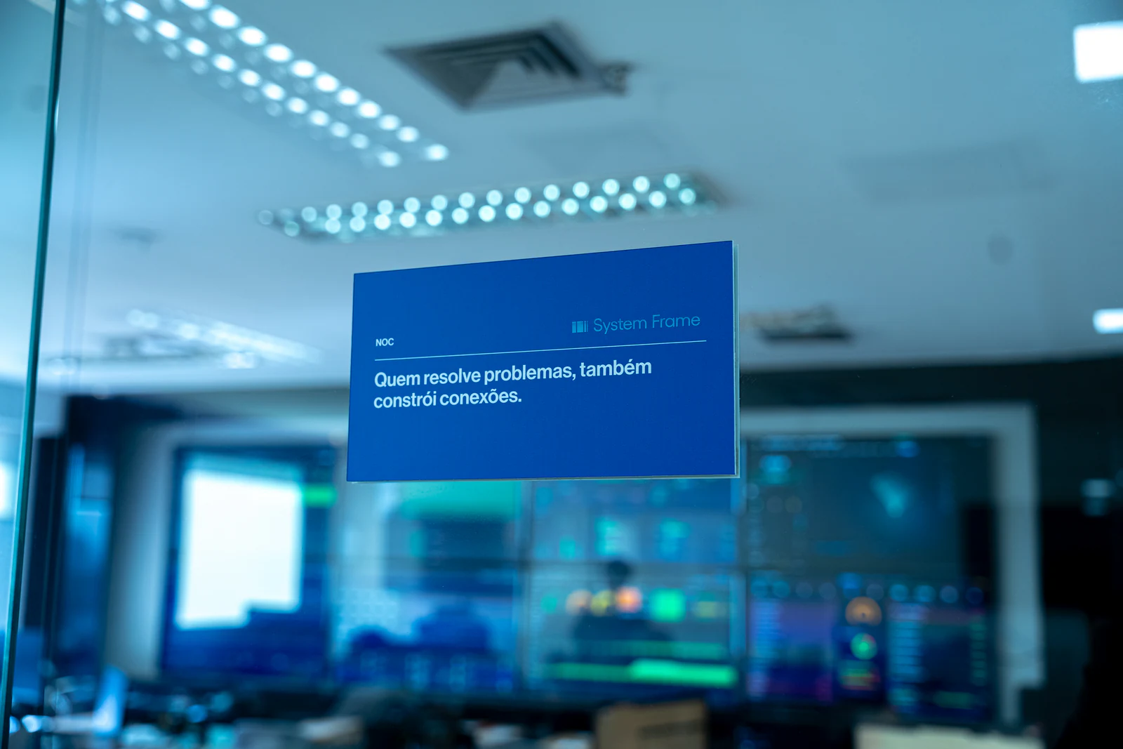 Placa de identificação do NOC da System Frame, com monitores desfocados ao fundo.